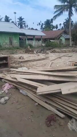 The video shows a scene of what appears to be a construction or demolition site in a tropical or subtropical location. There are piles of wooden planks and other debris scattered across the ground. In the background, there are houses, trees, and palm trees. Towards the end of the video, a small group of people can be seen standing near a house, and a person on a moped passes by. The overall impression is that of a disorganized and possibly damaged area.
