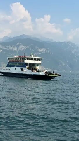 A ferryboat named "GRISALLO" sails across a body of water. The ferry is white and blue with a multi-level passenger cabin. People are visible on the upper decks of the ferry, some standing and others seated. A white car is parked on the lower deck of the ferry. The ferry is moving from left to right in the frame.

The background features a serene lake with gently rolling waves. Distant mountains rise majestically, their peaks partially obscured by a light haze. The sky is a clear blue with scattered, fluffy white clouds. The overall scene suggests a bright, sunny day, likely mid-morning or early afternoon. The setting appears to be a picturesque lakeside environment, consistent with the location context of Loveno, Italy. The activity is a ferry crossing, a common mode of transport for both locals and tourists in such areas, enabling the movement of people and vehicles across the water. The mood is peaceful and leisurely, evoking a sense of travel and natural beauty.