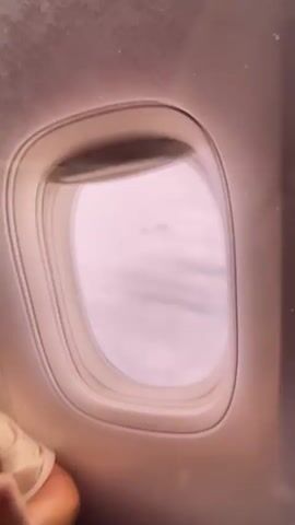 A video shows a view from an airplane window. The view is of clouds from above, with the wing of the plane visible. The sky is hazy and appears to be a light purple or pink color, suggesting either sunrise or sunset. The clouds are thick and layered, with some darker shadows visible between them, indicating depth and varying altitudes. The wing of the airplane is a light gray color and is angled across the frame, extending towards the right side of the image. There are some small water droplets visible on the inside of the airplane window, which distort the view slightly.