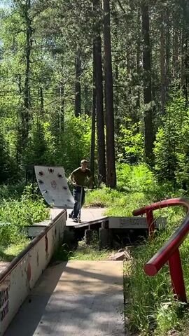 A young man on a scooter is performing tricks in a skatepark nestled within a forest. He is wearing a green t-shirt and blue jeans. The skatepark features a ramp with a spider graphic, a grind rail, and wooden platforms. Sunlight filters through the tall trees, indicating it is daytime and likely pleasant weather. The scene is natural and somewhat overgrown with lush green foliage surrounding the park. The man appears focused and determined as he maneuvers his scooter. There is another person visible in the background, also on a scooter. Text is visible on the side of one of the skatepark features, reading "BRICK COUNTRY."