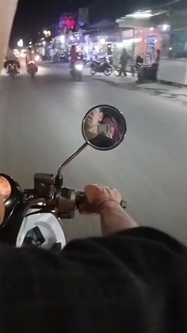 The video shows a first-person perspective of someone riding a white motorcycle at night on a street. The rider's hands are visible on the handlebars. A circular rearview mirror reflects the faces of two people, the rider and a passenger. The street is lined with buildings decorated with colorful lights and flags. Several other motorcycles and people are visible on the street. The rider is moving forward, and the scene is slightly blurry due to motion.