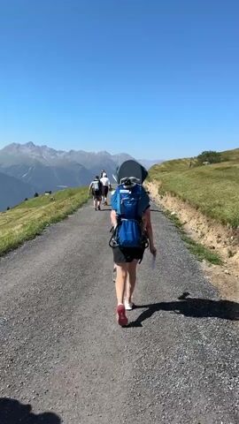 A group of people are walking up a gravel path on a mountainside. The person in the foreground is wearing a blue child carrier backpack. They are walking away from the camera, towards a distant mountain range. The sky is clear and blue. The path is lined with green grass and some bushes. The sun is casting shadows on the path. There is a slight incline on the path.