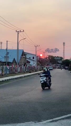 A motorcycle with two occupants rides down a road lined with buildings and trees. The sun is setting, casting a warm glow across the sky. The sky is a mix of pink and orange hues, with some clouds visible. The road is paved and appears to be in good condition. There are power lines running along the side of the road, and a cell tower can be seen in the distance. The buildings on the left side of the road appear to be houses or small businesses. Some of them are decorated with Indonesian flags. The motorcycle is white and has two people wearing helmets. The person in the back appears to be holding onto the person in the front. The motorcycle is moving away from the camera.
