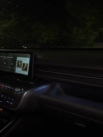 The video shows the interior of a car at night, facing forward. The car's dashboard and infotainment system are visible, with music playing on the screen. The song displayed is "Ceva si o Inima" by Mario Fresh. The car's interior lights are dim. Outside the car, it is dark, with trees and some distant lights visible through the windshield. There are faint reflections of raindrops on the windshield, suggesting it might be raining or has recently rained. The view outside changes slightly as the car appears to be moving or stationary with slight movement from the person holding the camera.