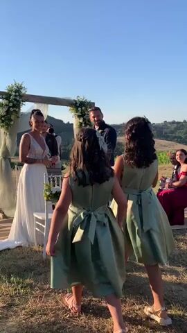 A wedding ceremony is taking place outdoors on a clear, sunny day. In the foreground, two young girls in matching light green dresses with sashes are walking away from the camera, towards the ceremony. They have dark hair and are wearing sandals. Behind them, a bride in a white wedding dress and a groom in a dark suit are standing under a wooden arch decorated with white flowers and green fabric. The bride is facing the groom and appears to be speaking to him. The groom is looking at the bride. To the right of the groom, there is a white chair decorated with white flowers and green ribbon. Further to the right, in the background, a woman in a burgundy dress is seated. The ceremony is set in a natural landscape with dry grass and rolling hills in the background. The sky is a clear blue. The video appears to be shot from a handheld perspective, with some slight movement.