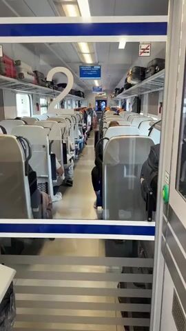 The video shows the interior of a train car, viewed from a doorway. Rows of empty white seats with silver accents are visible on either side of a central aisle. Luggage is stored on overhead shelves that run along the length of the car. Some passengers are seated in the chairs, mostly with their backs to the camera. The car is well-lit by overhead fluorescent lights. A large number "2" is prominently displayed on a shelf in the foreground, likely indicating the car number. A blue sign with white lettering is visible further down the aisle, possibly providing train information. The overall impression is a modern, clean train interior, likely in motion given the slightly blurred background and the presence of passengers.