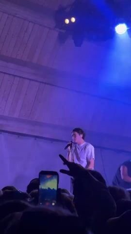 A man wearing glasses and a white t-shirt is singing into a microphone on a stage, illuminated by purple and blue stage lights. He is holding the microphone with his right hand. In front of the stage, a large crowd of people is gathered, many of whom are holding up their phones to record the performance. The stage has wooden beams and trusses supporting lights and speakers. The overall setting appears to be an outdoor concert or festival, given the presence of trees in the background and the open-air stage structure. The sky is dark, indicating it is nighttime. One person in the crowd is holding up a phone with a starry, cloudy image on its screen. The crowd is dense, with many individuals visible from the shoulders up. The atmosphere seems energetic and engaging, with the audience focused on the performer.