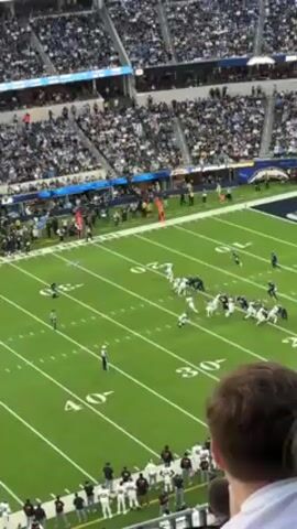 The video shows an American football game from an elevated perspective inside a stadium filled with spectators. The football teams, dressed in white and black uniforms, are lined up on a green field with yard lines. The play involves the offense attempting to move the ball forward, with players engaging in blocking and running. A fan is visible in the foreground, partly obscuring the view. The stadium's architecture is modern, and the overall atmosphere suggests an exciting live sports event.