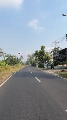 The video shows a paved road in a rural area. The road is empty and appears to be in good condition, with clear lane markings. On either side of the road, there are trees and vegetation. There is a clear blue sky with some white clouds. The sun is shining, indicating it is daytime. There are a few buildings visible in the distance, as well as utility poles and wires. A sign can be seen on the right side of the road. The perspective suggests the video is being recorded from a vehicle moving forward on the road, or from a person walking or riding a bike. There are no people or animals visible in the video. The overall impression is one of a peaceful and natural environment.
