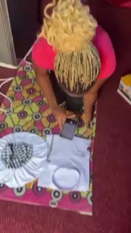 A woman is sitting on the floor taking pictures of items laid out on a colorful mat. She is wearing a pink t-shirt and black leggings, with her hair styled in blonde braids. On the mat are a white garment, a white cap, and a white and black checkered hat. The woman is holding a smartphone and appears to be reviewing the photos she has taken. The background includes a red curtain, a box, and other items suggesting an indoor setting.