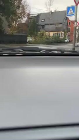 The video is taken from inside a car, showing the dashboard. Through the windshield, we see a suburban street corner. A house with a gray slate roof and yellow siding is visible, along with other houses in the background. Several parked cars are on the street, and a car crosses the intersection. The sky is overcast.