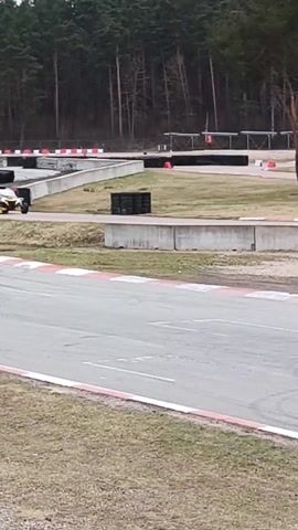The video shows an all-terrain vehicle driving on a race track. The vehicle is black and yellow, with two visible occupants. The race track is surrounded by low concrete barriers and has a white and red edge. Beyond the track, there is a grassy area and tall, bare trees. The weather seems overcast. The vehicle navigates around a curve on the track. A person can be seen standing in the background.