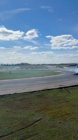 The video shows a race car drifting in a curve of a race track, on a sunny day with a blue sky with a few white clouds. The car is dark colored with green details and leaves a trail of white smoke behind as it moves. The race track is surrounded by green grass, and there's a light pole and buildings in the background. A person is standing on the grass near the track.