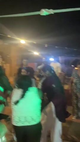 The video shows an outdoor nighttime gathering, possibly a party or celebration. Several people are visible, some dancing and interacting. Strings of lights are hung overhead, providing illumination. In the background, there are structures that appear to be made of natural materials like mud or adobe. The overall atmosphere is lively and social.
