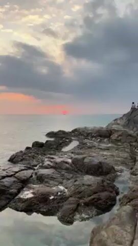 The video shows a pink-red sunset over the ocean. In the distance, a ship or boat is silhouetted against the setting sun. Rocky terrain is visible in the foreground, partially covered in small pools of water. The sky is cloudy with a mix of grey and soft pink hues. The water is calm, with small ripples reflecting the sunset. The camera zooms in slightly towards the sun and ship.
