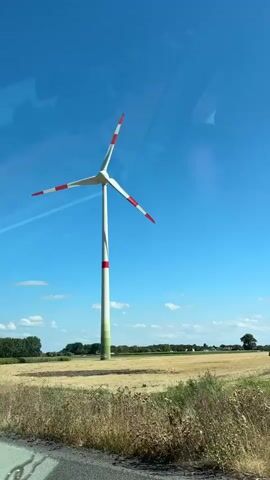The video shows a wind turbine in a field under a clear blue sky. The turbine has three white blades with red tips and a white and red striped tower. The field is dry and yellow, with some green foliage and trees in the background. The camera appears to be moving, possibly from inside a vehicle, as evidenced by reflections on the left side of the frame. The wind turbine is the central focus of the video, with the blades slowly rotating.