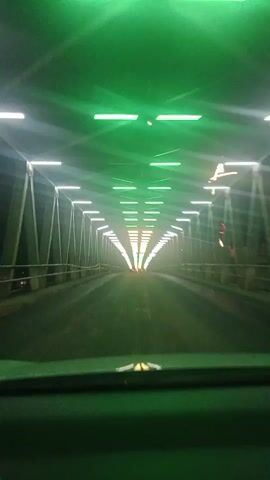 The video shows a point-of-view shot from inside a vehicle, looking forward through a tunnel or covered bridge. The structure features a metal framework with a series of lights on the ceiling. The lights are a mix of white and green, with the green lights concentrated at the beginning of the tunnel and the white lights extending into the distance. The road surface appears wet, and there is some atmospheric haze or fog. The tunnel narrows to a vanishing point in the distance, creating a sense of depth. The dashboard of the vehicle is partially visible in the foreground.