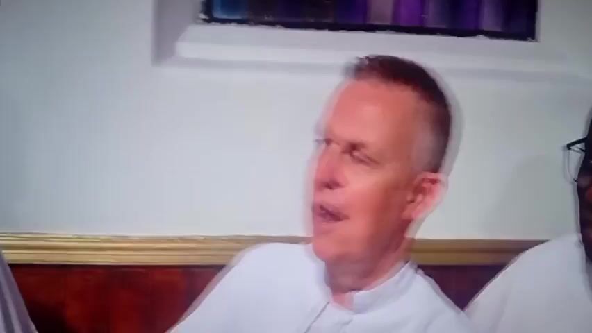 This media file captures a moment with two individuals, likely men, seated and engaged in conversation. The setting appears to be indoors, possibly a church or a formal gathering space, given the presence of what looks like a stained-glass window in the background and a decorative wooden paneling. The lighting suggests it's daytime.

The primary focus is on the man in the foreground, who is looking to his right and appears to be speaking. He has short, light-colored hair and is wearing a white garment, possibly a clerical robe or a uniform of some sort. His facial expression conveys animation, and his mouth is open as if he is mid-sentence. His right hand is slightly raised and clenched, suggesting emphasis or gesturing during his speech.

To his left, another man is partially visible. He is bald, wears dark-rimmed glasses, and is also wearing a similar white garment. His head is turned slightly away from the camera, and he seems to be listening or also speaking. His mouth is also open, indicating participation in the conversation.

The overall mood appears to be one of lively discussion or a sermon. There are no visible signs of the time of day or weather conditions. The location context provided, Ashipa, Nigeria, adds a cultural layer to the scene, suggesting it might be a religious or community event within that region. No text is visible in the media file.