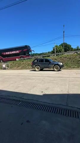 A red and black REGABUS tour bus is parked on an incline behind a dark gray Jeep. The Jeep has gold rims and appears to be off-road capable. A woman walks in front of the Jeep. The scene is outdoors with a clear blue sky. The Jeep is on a paved surface, and there is a manhole cover in the foreground.