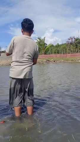 A young man stands in a body of water under a blue sky, preparing to cast a fishing net. He wears a light-colored t-shirt and dark shorts, with his bare legs submerged in the water. He holds a circular fishing net, adjusting it with both hands. In the background, there is land with some vegetation and structures.