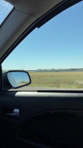 The video shows a view from inside a car, looking out the passenger window. The car is moving, as indicated by the reflection in the side mirror, which shows a road and other vehicles. Outside the window, there is a large, open field with dry grass. In the distance, there are several industrial buildings and structures, possibly part of an airport or industrial complex. The sky is clear and blue, suggesting a sunny day. The interior of the car, including the door panel, window frame, and a handle, is visible in the foreground.