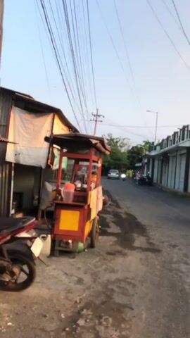 A food cart is parked on the side of a street, next to a building with a corrugated metal wall. A motorcycle is parked in front of the food cart. The street is paved with asphalt and has some debris on it. There are buildings on the other side of the street. Overhead, power lines crisscross the sky. The sky is clear and appears to be either sunrise or sunset. There are no people visible in the immediate vicinity of the food cart.