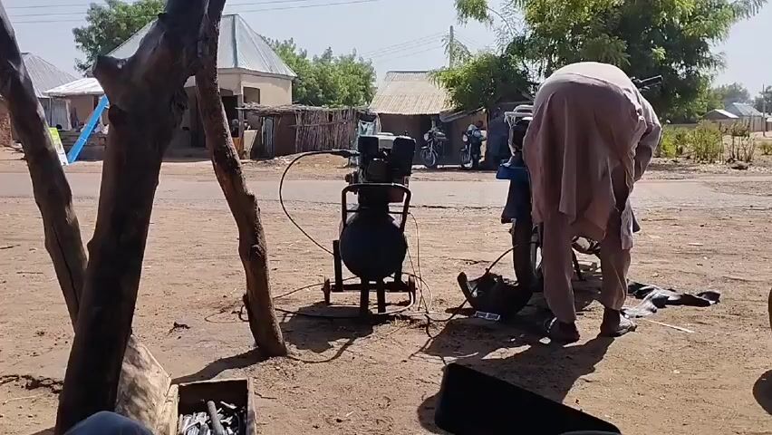 The video presents a daytime scene set in a rural environment. The foreground is shaded, dominated by a tree trunk to the left and a box of tools. In the center, a man wearing a light brown garment works on a motorcycle tire using a black air compressor. In the background, across a dirt road, are several houses and trees. Other individuals and motorcycles can be seen faintly in the distance. The scene appears to be a tire repair shop. A constant sound of machinery pervades the background.