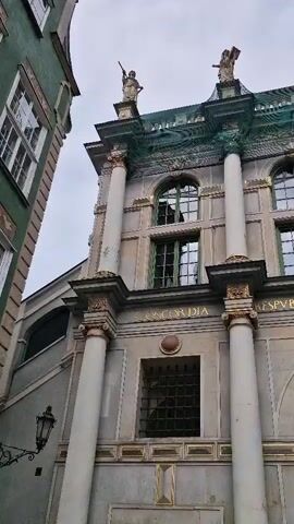 The video captures a low-angle view of the exterior of a historic building with classical architectural elements. The facade is adorned with columns, arched windows, and decorative sculptures at the top. Inscriptions are visible on the building's surface. Below the main facade, a large arched entryway leads to a glimpse of a brick structure and people walking. The sky appears overcast. The camera pans slightly, offering different perspectives of the building's facade.