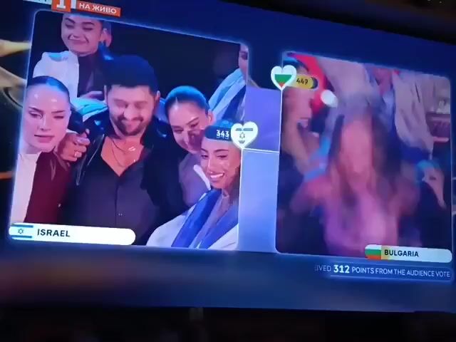 A video shows a split-screen display on a television. On the left, labeled "Israel", a group of people are holding an Israeli flag and smiling. The score 343 is displayed. On the right, labeled "Bulgaria", a group of people are jumping and cheering. The score 516 is displayed. The text "RECEIVED 312 POINTS FROM THE AUDIENCE VOTE" is shown below. It appears to be a live broadcast of a competition, possibly a singing or talent contest. The top left of the screen indicates the broadcast is live.