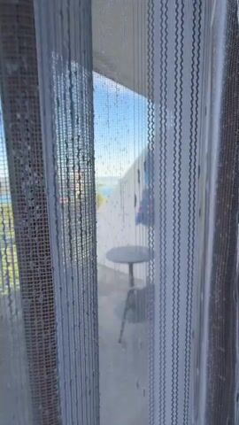 A hand pulls open a white, patterned curtain to reveal a balcony with a clear view of the water, green trees, and a red and white tower in the distance under a blue sky. The balcony is furnished with a gray table, two gray chairs, and a blue and red umbrella leaning against a white wall. A white railing lines the balcony edge. A road with a bus can be seen beyond the railing.