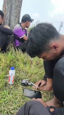 Four young men are sitting on the ground near a tree. Two of the men are looking at their phones. One man is holding a cigarette and appears to be lighting it. There is a bottle of water and a pack of cigarettes on the ground. The background shows a hilly, green landscape, possibly a tea plantation. The overall atmosphere appears casual.