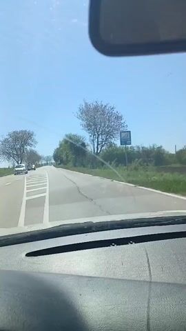 The video shows a car driving on a road. The road is paved and has white lines marking the lanes. There are trees and green vegetation on either side of the road. The sky is clear and blue. The camera is mounted on the dashboard of the car, and the video is taken from the driver's perspective.  The music is playing in the background.  There is a road sign visible.