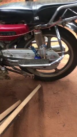 The video shows a red motorcycle, with silver accents, parked on a dirt surface in what appears to be a shed or lean-to structure with a corrugated metal roof. The motorcycle is facing left. In the foreground, there are wooden beams and plastic debris on the ground. The "DAYLONG" logo is visible on the side of the gas tank. The surrounding environment includes vegetation visible through the open sides of the structure.