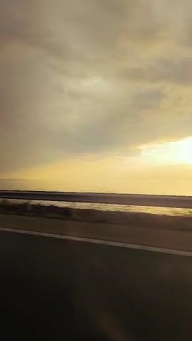 A video taken from a moving vehicle shows a sunset over the ocean. The sky is filled with clouds, and the sun is a bright, glowing orb on the horizon. The ocean is calm and reflective, with the sun's rays creating a shimmering path across its surface. A guardrail runs along the side of the road, separating it from the ocean. The road itself is dark and appears to be made of asphalt. The video is short and does not contain any people or significant actions.