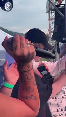 This video captures a lively outdoor event, likely a music festival, at Дрокино, Russia. The scene is set against a backdrop of a slightly overcast sky, suggesting it's late afternoon or early evening. In the foreground, a person is visible from the back, wearing a black t-shirt and a bright green wristband, indicating they are likely a participant or attendee. Their left hand, adorned with rings, is raised, possibly in a gesture of engagement or celebration.

Dominating the center of the frame is a DJ setup, featuring white Pioneer CDJs. A person wearing a black baseball cap and a white long-sleeved shirt is actively engaged with the DJ equipment. They are holding a microphone in their right hand, with intricate tattoos visible on their arm, including what appears to be a depiction of a jester and other stylized designs. This individual is also holding a smartphone in their left hand and a beverage can. Another person, possibly a friend or collaborator, is leaning over them, with their arm around the DJ's shoulder, also holding a can. The interaction between these individuals suggests a dynamic and possibly informal atmosphere.

Visible text includes "Pioneer" on the DJ equipment. While the full text on the can and smartphone is not clearly legible, the presence of a camera operator in the background suggests the event is being professionally documented. The overall mood appears energetic and celebratory, characteristic of a festival or concert setting. The lush green surroundings visible in the distance hint at a rural or semi-rural location.
