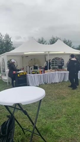 This video shows an outdoor event with several white tents set up on a grassy field. People are milling around the tents, and some are seated at tables. In the foreground, there is a white round table with a black trash bag underneath. In the background, there are trees and a cloudy sky. One tent appears to be a food stand, with food displayed on a table and people serving customers. Another tent has a sign that says "ШАУРМА". The overall atmosphere seems casual and festive.