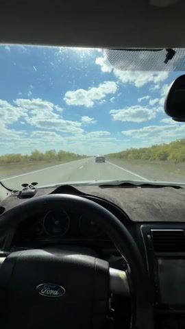 The video is filmed from the perspective of a driver inside a Ford vehicle, looking out the windshield. The car is on a highway with a white line marking the lanes. The sky is blue with fluffy white clouds, and the sun is shining brightly, creating a glare on the windshield. Trees and foliage line the sides of the road, and another white car is visible in the distance on the highway ahead. The steering wheel, dashboard, and part of the car's interior are visible in the foreground. The video captures the experience of driving on a sunny day.