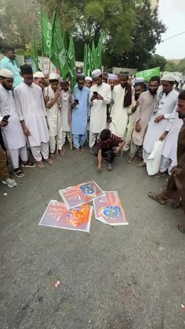 A group of people, mostly young men wearing white Islamic attire and skullcaps, are gathered in a circle on a paved street. They are holding up green flags with white writing. In the center of the circle, a man in a striped shirt is kneeling and appears to be burning several documents or posters. Flames and smoke are visible, and the people are watching the burning. The environment appears to be outdoors, with trees and buildings in the background. The sky is overcast.