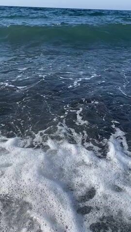 The video shows a close-up view of waves crashing on a dark, pebbly beach. The water is a deep blue, and the foam from the waves creates a white pattern on the dark rocks. The camera is positioned very low, near the water's edge, giving the viewer a sense of being right in the action. In the background, the ocean stretches out to the horizon under a clear sky. The lighting suggests it is daytime. There are no people or objects visible in the video other than the natural elements of the beach and the sea.