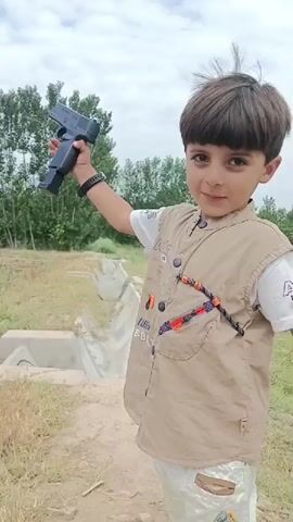 A young boy with dark hair stands outdoors in a grassy area with trees in the background. He is wearing a white t-shirt, a tan vest with decorative beads and buttons, and patterned pants. The boy is holding a black toy gun in his right hand, raising it slightly. He is looking towards the camera with a smile. The video captures him in a series of still frames, with a slight pause between each.