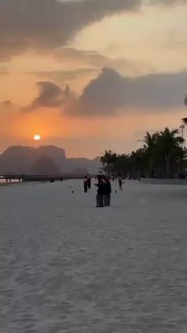 The scene is a beach at sunset. The sky is filled with orange hues from the setting sun, partially obscured by clouds. In the background, there are mountain silhouettes. The beach itself is sandy and relatively flat. A row of palm trees lines the edge of the beach. Several people are present on the beach, including a pair standing closer to the viewer, seemingly observing the sunset or taking pictures. Some other individuals are walking in the distance. The lighting creates a warm and serene atmosphere.