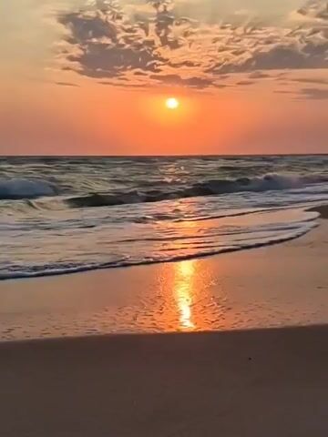 This is a slow-motion video of a beach at sunset. The sun is a bright orange orb near the horizon, casting a warm glow across the sky, which is filled with wispy clouds. The ocean is calm, with gentle waves rolling onto the sandy shore. The wet sand reflects the colors of the sunset. There are no people or objects visible in the video.