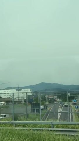 The video pans across a grey, overcast sky, revealing a scene from a moving vehicle. In the foreground, green grass and weeds line a roadside. Beyond the grass, a highway with several cars and a bus is visible. A large white building with "AUTOPARKING" written on it stands in the middle ground. Further in the background, mountains rise against the cloudy sky. The video continues to pan, showing a parking lot filled with garbage, including several garbage trucks, discarded items, and litter. The overall impression is one of urban sprawl and neglect.