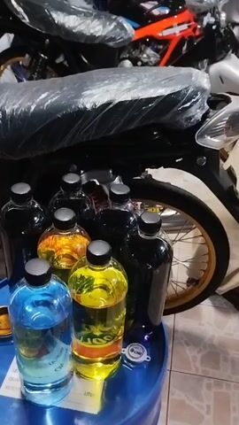 The media file shows a collection of bottles filled with colorful liquids, predominantly blues, yellows, and blacks. These bottles are arranged on a blue tray, which appears to be sitting on a larger blue container, possibly a barrel. In the background, several motorcycles are visible, some of them covered in plastic. The setting seems to be a workshop or a storage area for motorcycle parts or accessories, given the presence of the motorcycles and the arrangement of items. The lighting suggests it might be daytime, but there are no clear indicators of weather or specific time of day. The overall mood is neutral, focusing on the presentation of the bottles and the motorcycles. Some text is visible on the blue tray and possibly on the bottles, but it's mostly illegible due to the low resolution and angle. The location context provided is Si Sunthon, Thailand.