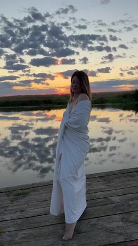 The video shows a woman standing on a wooden dock by a body of water. She is wrapped in a white blanket, and her shoulders are bare. The sun is setting, casting an orange glow across the sky and reflecting on the water. The sky is filled with clouds, which are also lit by the setting sun. The woman is looking off to the side with her eyes closed, and she appears to be smiling. The overall mood of the video is peaceful and serene.