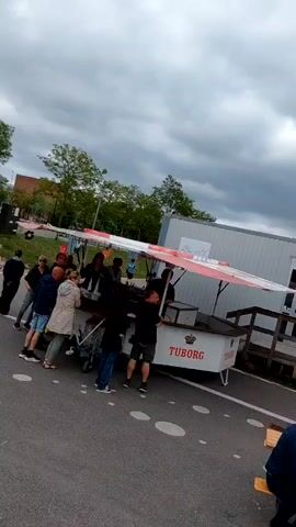 The video depicts an outdoor event on a cloudy day. Several people are gathered around food and drink vendors, including one selling Tuborg beer. Some individuals are seated at picnic tables, eating and socializing. Other people are standing in line at the vendors or walking around the area. There are white tents set up nearby, suggesting an organized event. The surrounding area includes buildings, trees, and a brick wall.