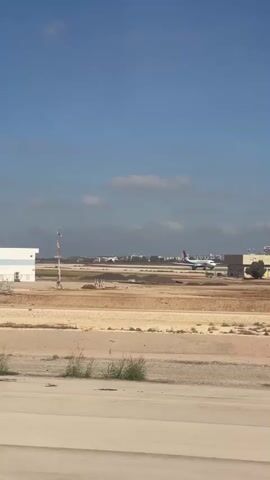 This is a video clip of an airplane at an airport. The camera is positioned inside an airplane, looking out the window. The perspective shows the wing of the plane in the foreground on the left side of the frame. The wing is white with a pink stripe at the tip, and the text "wizzair.com" is visible on the stripe. In the background, across a sandy airfield, there are several airport buildings and some distant city structures. An airplane with a red and blue tail is visible on a runway in the middle distance. The sky is blue with scattered clouds. The video shows the airplane taxiing on the ground.