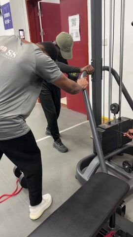 Two men are in a gym, one using a lat pulldown machine. One man is wearing a light gray shirt with the word "BOSS" on it, black pants, and white shoes; he is bent over slightly, facing away from the camera. The other man wears a long-sleeved black shirt, black pants, gray shoes, and a light green hat. He is holding onto the lat pulldown machine in front of the first man. The machine is gray and black with a seat, a metal bar for pulling, and weight plates stacked up. There is a red cord on the floor near some black dumbbells. A red door and a sign are visible in the background. The floor is gray.