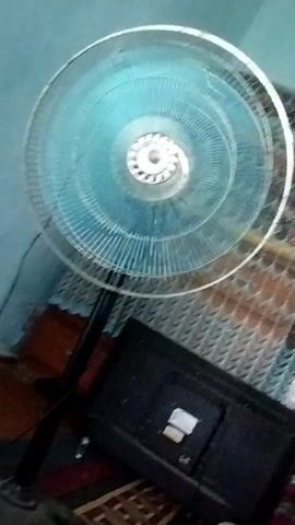 A standing fan with a transparent blue fan guard and a black stand is positioned behind a black television set. The fan is in motion, with its blades rotating. The fan is on a dark carpeted floor, and behind it, a patterned curtain can be seen. The room appears to be dimly lit.