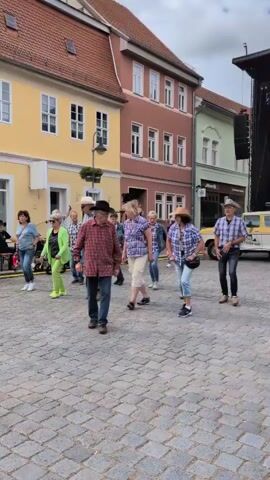 The video shows a group of people, primarily adults, engaged in a line dance outdoors on a cobblestone square. They are dressed in casual clothing, with many wearing plaid shirts and some sporting cowboy hats, suggesting a country or Western theme. The dancers are arranged in two rows, facing the same direction and performing synchronized movements. In the background, there's a stage with a band playing, a vintage pickup truck, and buildings with red tile roofs. The sky is overcast. The overall atmosphere appears to be festive, possibly a street fair or community event.